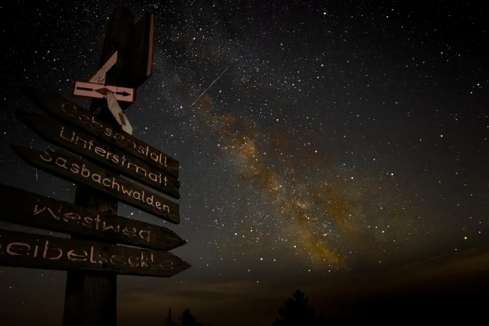 Wegweiser mit mehreren Schildern vor einem klaren Nachthimmel mit sichtbarer Milchstraße und zahlreichen Sternen über der Hornisgrinde im Schwarzwald.