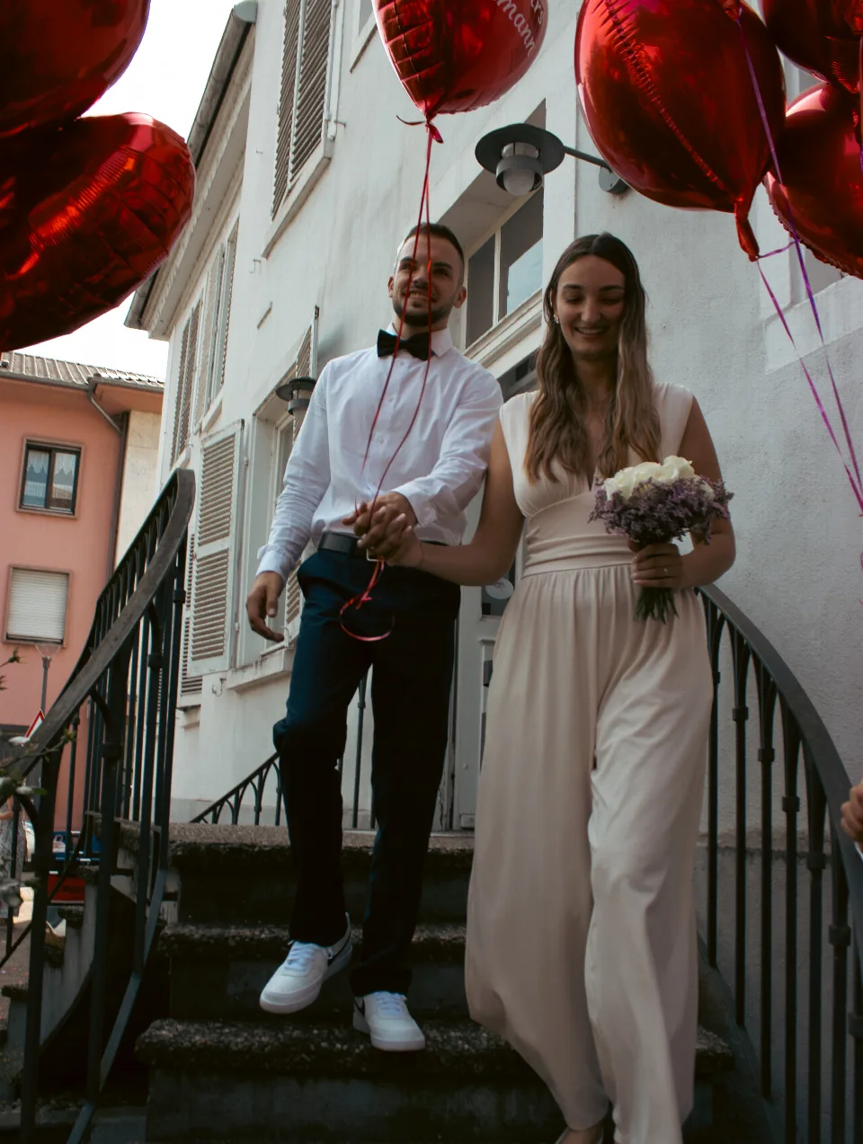 Brautpaar in festlicher Kleidung steigt Hand in Hand eine Außentreppe am weißen Standesamtsgebäude hinab. Die Frau trägt ein langes Kleid und einen Blumenstrauß, der Mann ein Hemd mit Fliege und weiße Schuhe. Rote Herzluftballons sind im Hintergrund zu sehen.