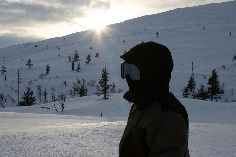 Silhouette einer Person mit Skibrille und Kapuze vor einem verschneiten Hügel im Pallas-Yllästunturi-Nationalpark bei Sonnenuntergang.