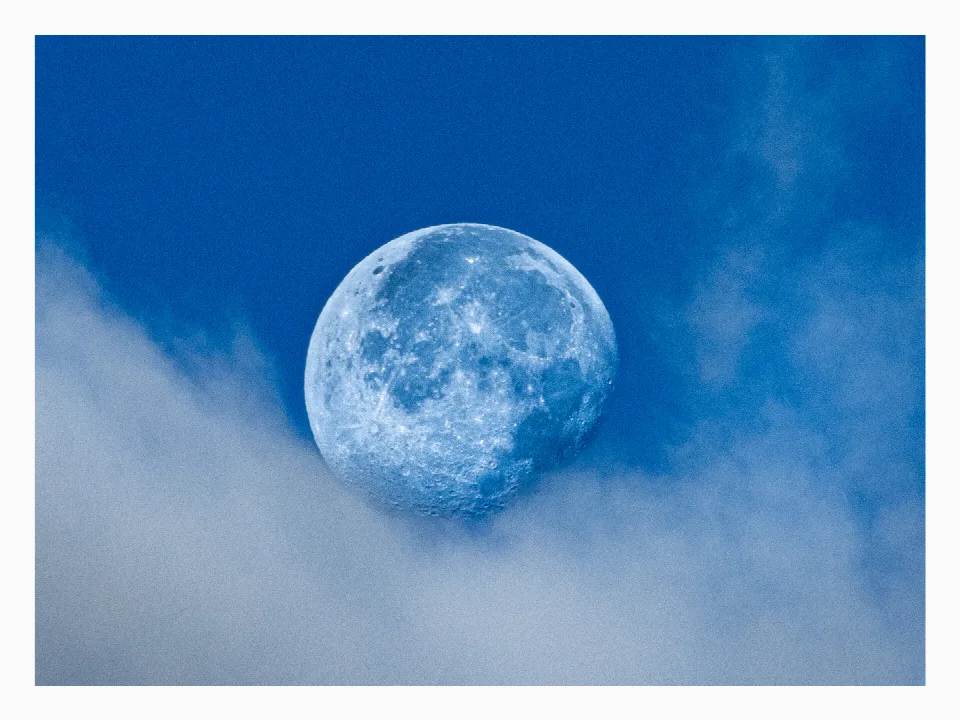 Zunehmender Mond am blauen Tageshimmel, umgeben von weißen Wolken.