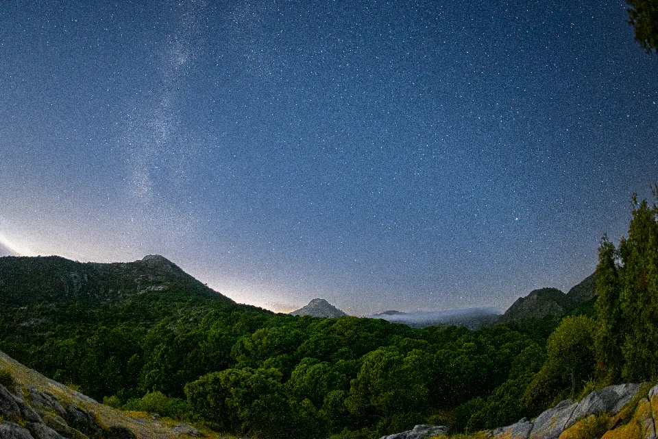 Sternenhimmel mit sichtbarer Milchstraße über bewaldeten Bergen, mit Felsen im Vordergrund am Mirador del Corzo in Málaga, Spanien.
