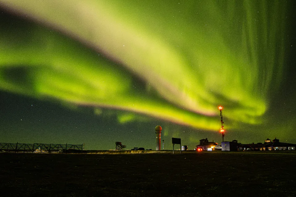 Grüne Nordlichter am Nachthimmel über der Kaunispään-Sternwarte in Finnland, mit beleuchteten Gebäuden und Antennen im Vordergrund.